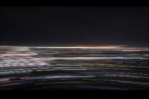 The abstract video still shows a long-exposure photography. A horizontal, fast movement creates coloured lines against a dark background in the shape of a rainbow. It invites associations, such as of a city at night with lights and traffic.