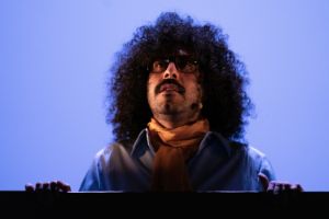 We see a close-up of a person against a blue illuminated background, that has long black curls, wears glasses and looks up to the stage sky.