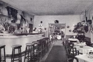 A still deserted, tidy bar with many plants and wall decorations. In the front on the left: a counter with bar stools. On the right: tables with place settings. An opening to the next room reveals instruments waiting for the band.