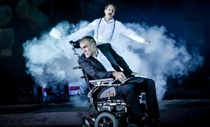 In einer Szene der Inszenierung „Reich des Todes“ sitzt der Schauspieler Sebastian Blomberg in einem elektrischen Rollstuhl, hinter ihm steht der Schauspieler Holger Stockhaus in einer Nebelwolke.