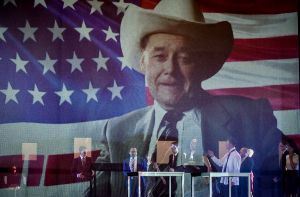 In einer Szene der Inszenierung “Reich des Todes” ist der Schauspieler Wolfgang Pregler mit einem Cowboyhut vor der US-amerikanischen Flagge in einer Projektion zu sehen.  