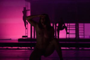 A naked woman screams on a stage bathed in purple light. Behind her at a water tank are other unclothed women.