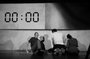 Drei Personen erheben sich vom Boden. Eine Uhr auf einer Leinwand zeigt 00:00.