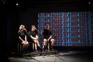 Eine Frau und zwei Männer sitzen an Mikrofonständern auf einer Bühne, mit dem Titel der Performance „PUTINPROZESS“ auf eine Leinwand im Hintergrund projiziert.