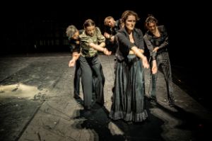 Six performers in dark costumes hold their right arm with their left hand.