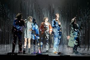 Six people at the edge of the stage in front of numerous poles attached to the floor