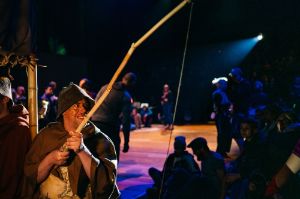 A person in a brown cloak holds a stick with a string attached to it