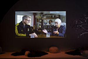 A screen shows two people talking in a living room