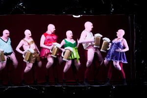 An ensemble with costumes reminiscent of sex dolls is on a black stage.