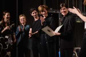 Gisèle Vienne holds the Theatertreffen certificate in her hand. People stand around her and clap.