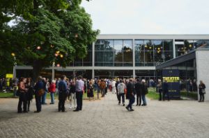 The audience enjoys the pleasant summer evening in front of the Berlin Festival House. On the left is a large tree, in the background is the building, on the right is a stretched poster with the Theatertreffen logo, people are standing together everywhere.