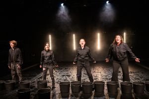 Four black-clad people stand with open mouths behind black buckets on a stage.