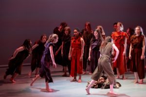 A group of young people stand on a stage with serious expressions on their faces and perform a circular movement. In the centre there are clothes on the floor.