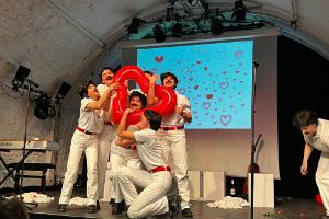 Seven people in white clothing with glued-on moustaches sing fervently on stage. Together they hold a red inflatable plastic heart in front of them.