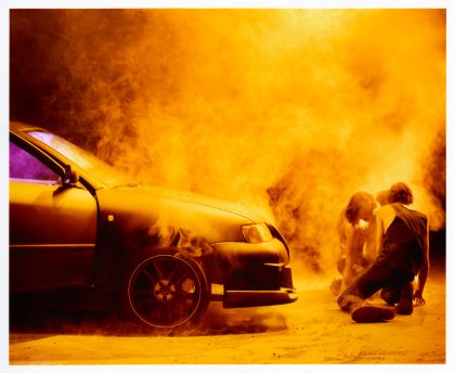 Two performers kneel on stage in front of a car, it is foggy around them.
